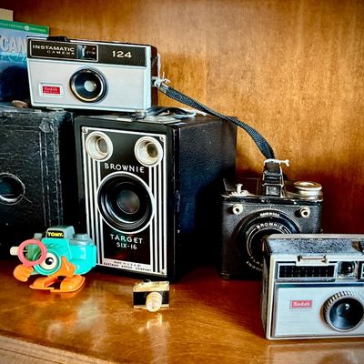 Sale Photo Thumbnail #191: Collection of vintage cameras, including a Kodak Brownie Target Six-16 box camera, a Kodak Instamatic 124, and a Kodak Instamatic 100, displayed alongside a small Tomy wind-up toy.