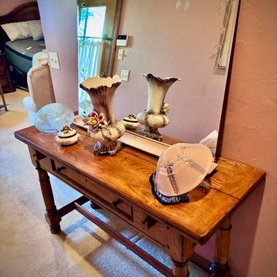 Sale Photo Thumbnail #124: Rustic wooden console table with drawers, displaying a large frameless mirror with etched floral details, a decorative ceramic vase, and various glassware pieces.