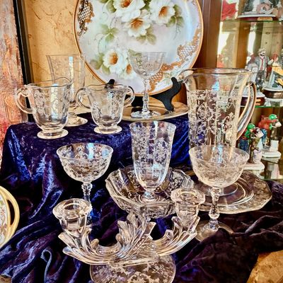 Sale Photo Thumbnail #25: Assorted elegant etched glassware, including a pitcher, goblets, and a double candlestick holder, displayed alongside a hand-painted floral porcelain plate.