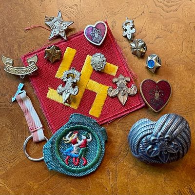 Sale Photo Thumbnail #89: Collection of vintage Boy Scouts of America memorabilia, including a red and yellow swastika patch (used as a symbol of good luck prior to WWII), various metal pins, merit badges, and a metal neckerchief slide.