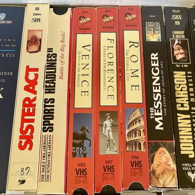 Sale Photo Thumbnail #92: Collection of vintage VHS tapes including "JFK Director's Cut," "Sister Act," and several Johnny Carson compilations, showing some wear on the cardboard sleeves.