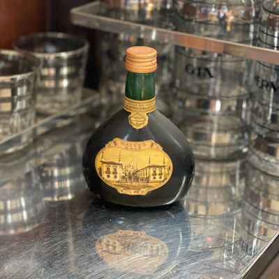 Sale Photo Thumbnail #157: Vintage Mateus Rosé wine bottle with its signature flask shape and original label, displayed on a mirrored bar cart alongside glassware.