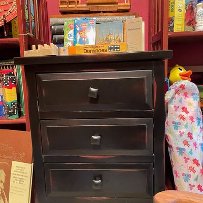 Sale Photo Thumbnail #193: Three-drawer black wooden nightstand with a distressed finish and square metal knobs, styled with vintage games and books on top.