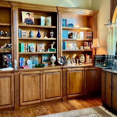 Sale Photo Thumbnail #30: Large three-section wooden library bookcase unit with lower cabinets, filled with books, collectibles, and vintage toys, alongside an Art Deco style bar cabinet.