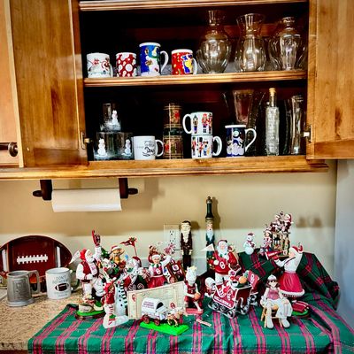 Sale Photo Thumbnail #200: Collection of University of Oklahoma Sooners themed Christmas decorations, including Santa figurines, a sleigh, and mugs, displayed alongside holiday glassware in a wooden cabinet.