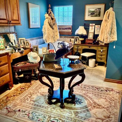 Sale Photo Thumbnail #35: Vintage dressing room display featuring a dark wood octagonal accent table, a French provincial style vanity desk, fur coats, and various antique accessories on a floral patterned area rug.