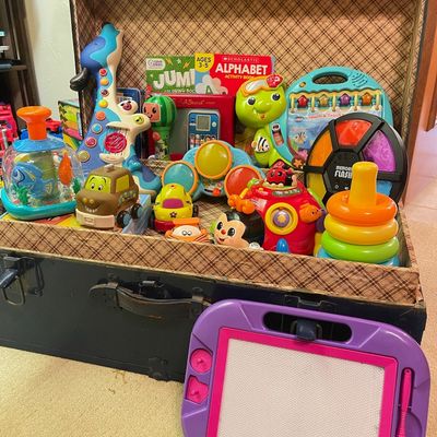 Sale Photo Thumbnail #109: Assortment of colorful children's toys and activity books displayed inside a vintage plaid-lined trunk, with a purple magnetic drawing board in the foreground.