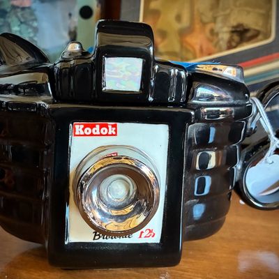 Sale Photo Thumbnail #47: Vintage ceramic teapot shaped like a Kodak Brownie camera, featuring a glossy black finish and metallic silver details.