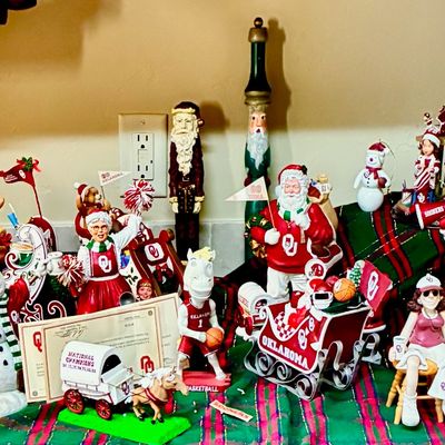 Sale Photo Thumbnail #203: Collection of University of Oklahoma Sooners themed Christmas figurines, including Santas, cheerleaders, and a sleigh, displayed on a plaid runner.
