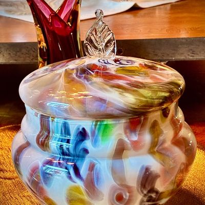 Sale Photo Thumbnail #205: Vintage hand-blown art glass lidded jar featuring a vibrant, multi-colored swirl pattern on a white base, topped with a clear glass leaf finial.