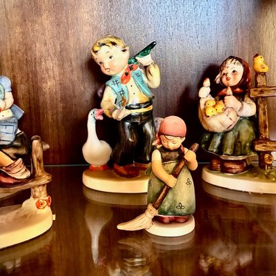 Sale Photo Thumbnail #76: Collection of vintage Goebel Hummel figurines, featuring children in traditional attire with animals and rustic elements, displayed on a glass shelf.