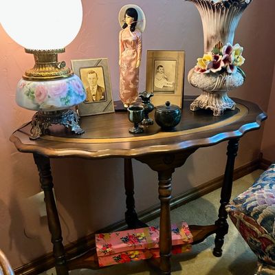 Sale Photo Thumbnail #101: Vintage wooden console table with scalloped edges and turned legs, displaying a collection of decorative items including a hand-painted hurricane lamp, framed vintage photographs, a doll, and an ornate ceramic vase.