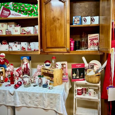 Sale Photo Thumbnail #202: Collection of University of Oklahoma Sooners memorabilia, including dolls, mugs, ornaments, and kitchenware, displayed on a table and in wooden cabinets.