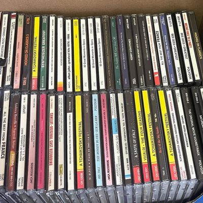 Sale Photo Thumbnail #236: A cardboard box filled with a large collection of vintage CDs, featuring various genres including classical, pop, and movie soundtracks.