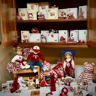 Sale Photo Thumbnail #188: Collection of University of Oklahoma Sooners memorabilia, featuring collegiate porcelain dolls, plush bears, holiday ornaments with original boxes, and various crimson and cream themed collectibles.