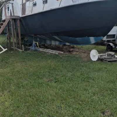 Vintage cabin cruiser boat on jack stands in a grassy yard, showing a dark hull and white upper deck in need of restoration.