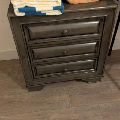 Sale Photo Thumbnail #26: Modern three-drawer nightstand in a gray wood finish with metallic knob pulls, topped with folded towels.