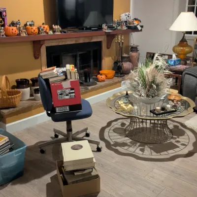 Sale Photo Thumbnail #11: Living room filled with various items for sale, including a mid-century wire-base coffee table with a glass top, a blue office chair, books, and Halloween decorations.