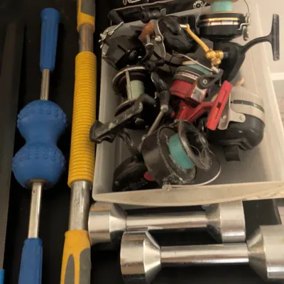 Sale Photo Thumbnail #40: Assorted sporting goods and tools, including a Rawlings baseball glove, a bin of fishing reels, a pair of chrome dumbbells, a yellow-handled hammer, and a blue massage roller bar.