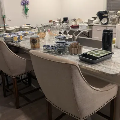 Sale Photo Thumbnail #9: Pair of contemporary upholstered bar stools with nailhead trim, shown alongside an assortment of kitchenware, glassware, and small appliances displayed on a kitchen island.