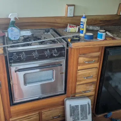 Sale Photo Thumbnail #97: Vintage stainless steel marine stove and oven unit built into wooden cabinetry, alongside a compact refrigerator and various boat maintenance supplies.