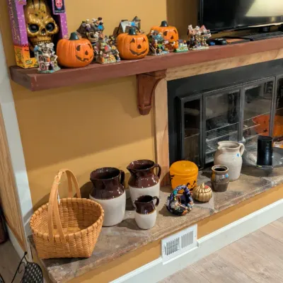 Sale Photo Thumbnail #22: Assorted Halloween decorations, including jack-o'-lanterns and a skull plaque, displayed on a mantel above a hearth featuring stoneware jugs and a woven basket.