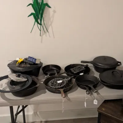 Sale Photo Thumbnail #12: Collection of vintage cast iron cookware, including skillets, Dutch ovens, and lids, displayed on a folding table beneath a metal iris wall sculpture.