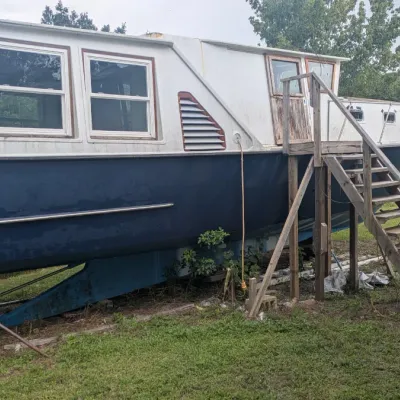 Sale Photo Thumbnail #95: Vintage houseboat on a dry dock, featuring a blue hull, white upper cabin with residential-style windows, and a wooden access staircase.
