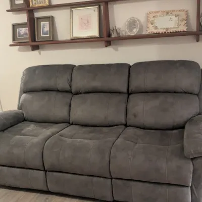 Sale Photo Thumbnail #38: Contemporary three-seat reclining sofa upholstered in dark gray microfiber fabric, positioned beneath a wall-mounted wooden shelving unit.