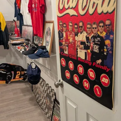 Sale Photo Thumbnail #74: Vintage Coca-Cola Racing Family poster taped to a door, alongside various NASCAR memorabilia including hats, shirts, and framed prints.