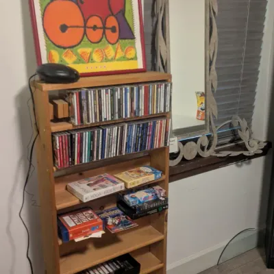 Sale Photo Thumbnail #84: Tall pine media tower filled with CDs, cassette tapes, and board games, topped with a colorful framed train print, alongside a decorative scrollwork wall mirror.