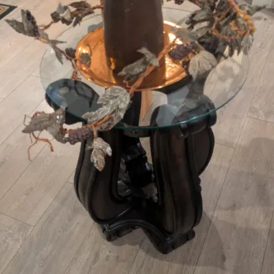 Sale Photo Thumbnail #72: Round glass-top accent table featuring a dark carved wood pedestal base, styled with a copper tray, a wooden vase, and a decorative faux leaf garland.