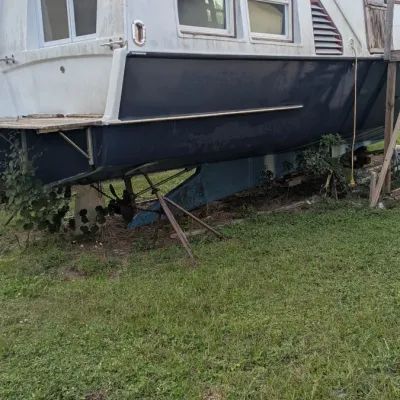 Vintage fiberglass houseboat on a trailer, featuring a white cabin over a dark blue hull, in need of restoration.