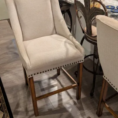 Sale Photo Thumbnail #13: Upholstered counter-height barstool with beige fabric, nailhead trim, and wooden legs, shown alongside other barstools and kitchenware.