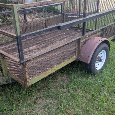 Sale Photo Thumbnail #47: Single-axle utility trailer with mesh sides, a wooden deck, and a rear ramp, showing some rust and wear from outdoor use.