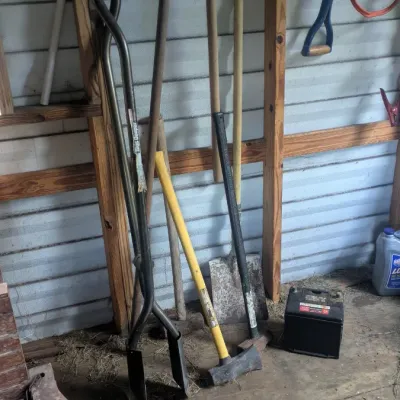 Sale Photo Thumbnail #46: Assorted lawn and garden tools, including a post hole digger, splitting maul, and shovels, leaning against a shed wall.