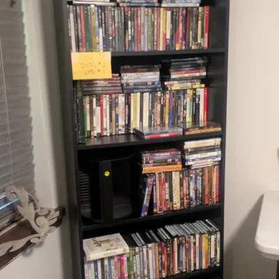 Sale Photo Thumbnail #83: Tall black laminate five-shelf bookcase filled with a large collection of DVDs and VHS tapes, topped with decorative wooden duck decoys.