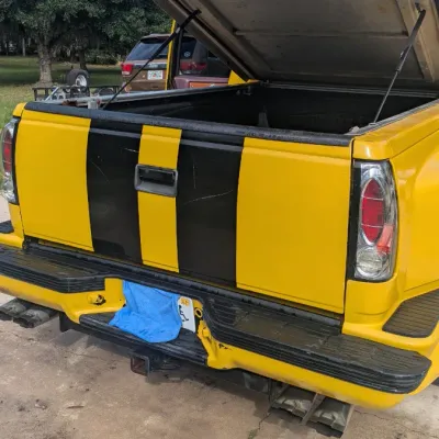 Sale Photo Thumbnail #57: Yellow pickup truck bed with black racing stripes, featuring a raised hard tonneau cover and dual exhaust.