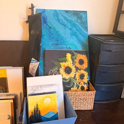 Sale Photo Thumbnail #112: Assorted amateur acrylic paintings on canvas, including a large blue fluid art piece, a sunflower painting, and a small sunset landscape, displayed alongside art supplies.