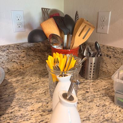Sale Photo Thumbnail #141: Assorted kitchen utensils including wooden spoons, spatulas, and yellow-handled flatware, displayed in various countertop holders alongside white ceramic oil and vinegar dispensers.