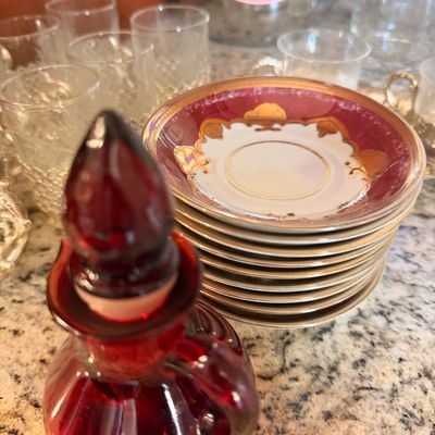Sale Photo Thumbnail #146: Vintage ruby red glass decanter with stopper, shown alongside a stack of porcelain saucers featuring a maroon and gold gilt border.