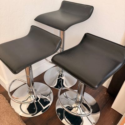 Sale Photo Thumbnail #4: Trio of modern adjustable barstools featuring black faux leather seats, chrome-finished pedestals, and integrated footrests.