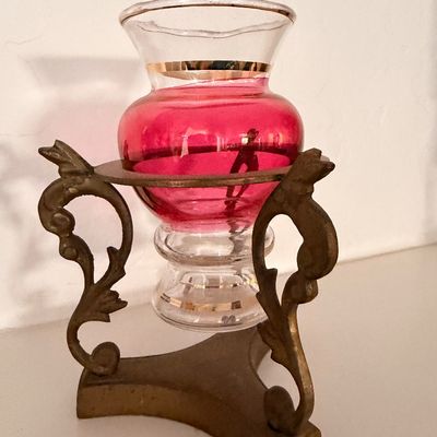 Sale Photo Thumbnail #15: Vintage cranberry glass urn-style vase with gold trim, resting in an ornate brass-plated metal stand.
