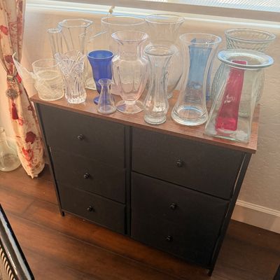 Sale Photo Thumbnail #87: Collection of assorted glass vases in various shapes, sizes, and colors, including clear, blue, and pink, displayed on a modern black five-drawer storage chest.
