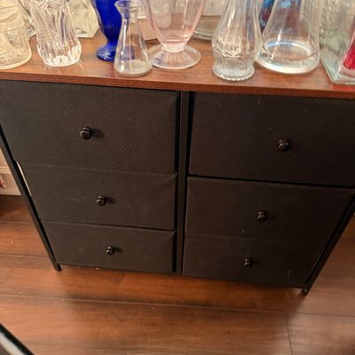 Sale Photo Thumbnail #65: Modern six-drawer storage chest with black fabric bins, a dark metal frame, and a wood-look top, shown holding a collection of glass vases.