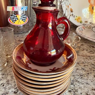 Sale Photo Thumbnail #83: Vintage ruby red glass decanter with matching stopper, displayed on a stack of porcelain saucers with gold trim.