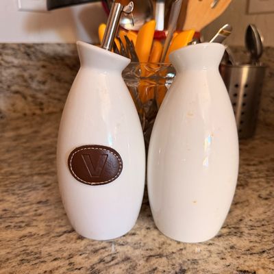 Sale Photo Thumbnail #75: Pair of white ceramic oil and vinegar cruets with metal pour spouts, one featuring a brown leather patch with the letter 'V'.