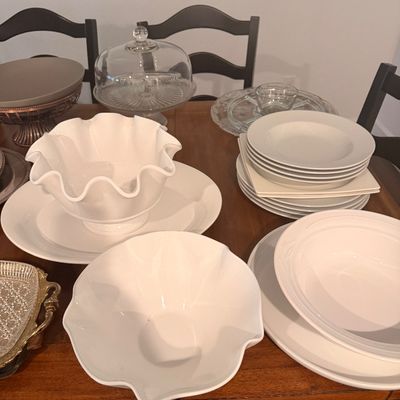 Sale Photo Thumbnail #97: Assorted white ceramic tableware, including ruffled serving bowls and stacked plates, displayed alongside glass serving pieces on a wooden dining table.