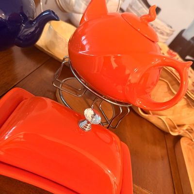 Sale Photo Thumbnail #94: Vibrant orange ceramic teapot on a wire stand, paired with a matching covered butter dish featuring a clear faceted knob.