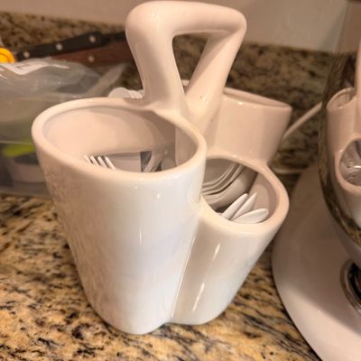 Sale Photo Thumbnail #142: White ceramic flatware caddy with a central carrying handle, holding plastic utensils on a granite countertop.